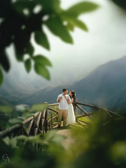 Framed by natural foliage, the couple shares a quiet moment on a wooden bridge overlooking the valley. This composition technique adds depth and intimacy to this scenic pre-wedding photograph.