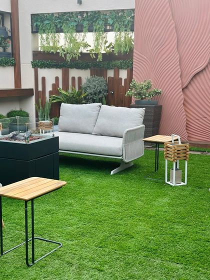 A modern terrace designed for comfort and style. This space features a firepit, sleek grey sofa, and a stunning textured accent wall, all surrounded by artificial turf and layered planters for a complete look.