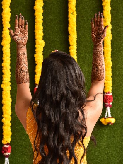 A beautiful back shot of a bride, her arms raised to display the full extent of her bridal mehendi. The design complements her festive yellow attire perfectly.