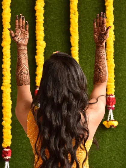 A beautiful back shot of a bride, her arms raised to display the full extent of her bridal mehendi. The design complements her festive yellow attire perfectly.