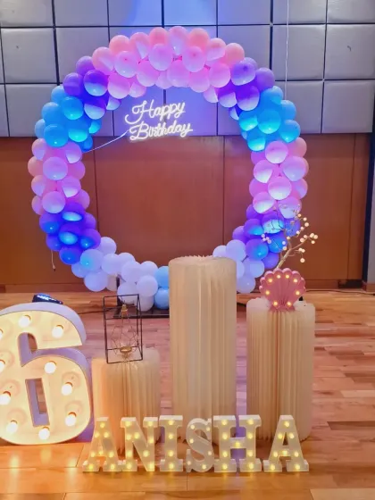 A colorful setup for a 6th birthday, featuring a full ring of pastel and blue balloons, a neon sign, and a large light-up number '6'.