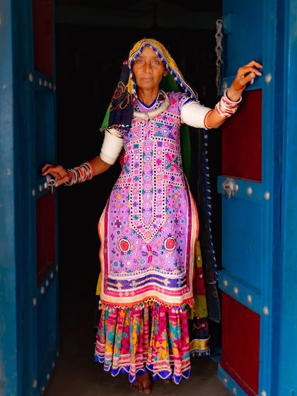 A Meghwal woman in a stunningly embroidered purple dress stands in the doorway of her home. The vibrant colors of her attire and the blue door create a beautiful composition.
