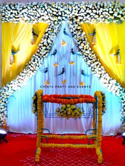 A traditional naming ceremony stage with a flower-decorated cradle. The backdrop features yellow and white drapes framed by a floral arch.