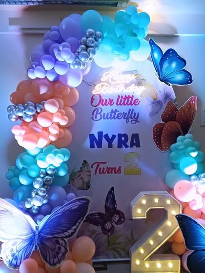 A 'Little Butterfly' theme for Nyra's 2nd birthday. The backdrop is adorned with large, realistic butterfly cutouts and a colorful balloon arch, all illuminated for a magical effect.