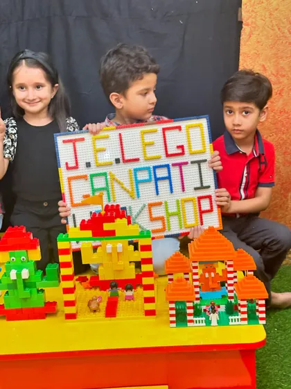 Three children gather behind their LEGO creations, including a sign that reads "J.E. LEGO GANPATI WORKSHOP," celebrating their collaborative effort.