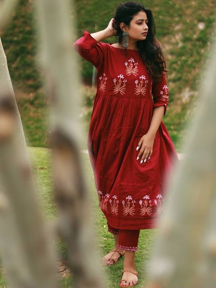 An outdoor fashion portrait taken in a park in Jaipur. The model stands confidently in a beautiful red embroidered kurta set, with the trees providing a natural, slightly obscured frame that adds depth to the composition.