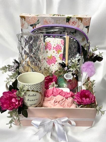 A lovely pamper hamper for her in a floral box. It includes a crystal-cut vase, a patterned mug, a clear pouch with beauty items, and is decorated with pink flowers.