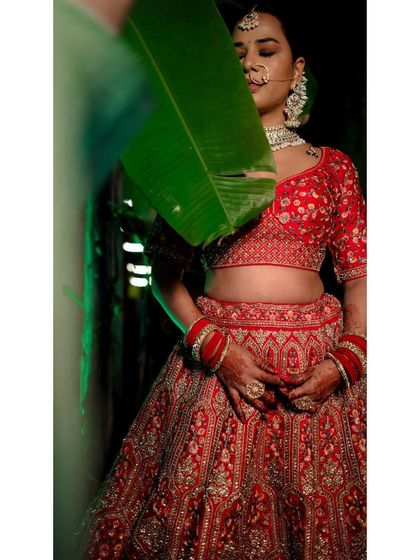 An artistic bridal portrait with a large green leaf partially covering her. The vibrant red of her lehenga contrasts beautifully with the green, creating a unique shot.