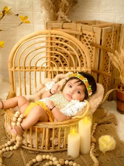 A baby girl lies in a wicker peacock chair, surrounded by yellow flowers and rustic props.