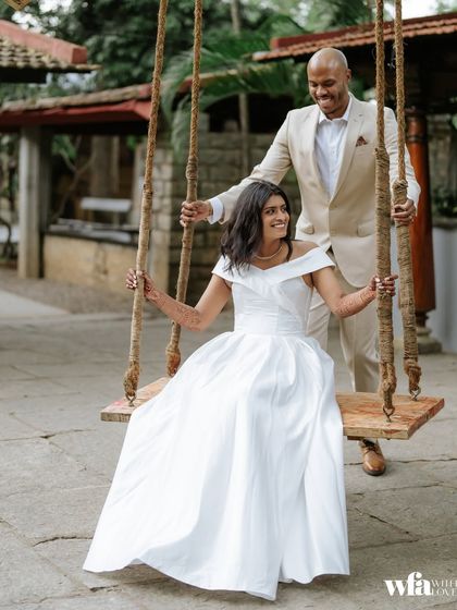 A modern couple, the groom in a suit and the bride in a white gown, share a moment on the rustic wooden swing.