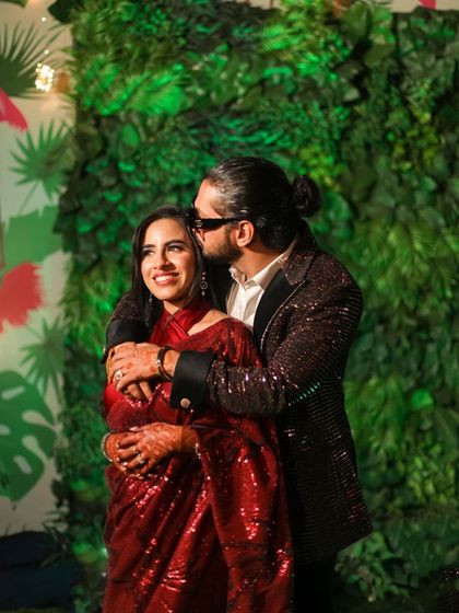 A candid, affectionate moment from an engagement party. The groom embraces the bride from behind and kisses her cheek, a spontaneous display of love captured against a vibrant green backdrop.
