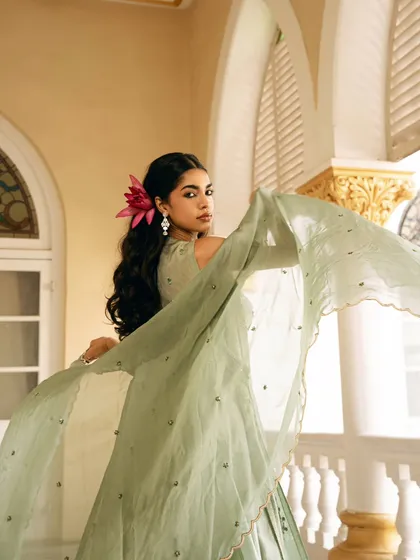 The movement of the dupatta is frozen in this shot, adding a sense of life and grace to the portrait. The flower in the model's hair complements the soft green of the ethnic outfit.