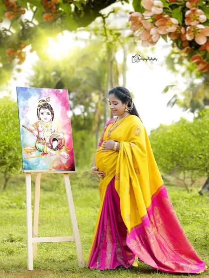 A creative outdoor portrait of a mother-to-be in a yellow and pink saree, looking at a painting of Baby Krishna.