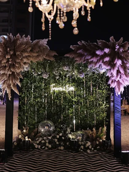 A stylish photo booth with a "Forever & Ever" neon sign on a green wall, framed by pampas grass and disco balls.