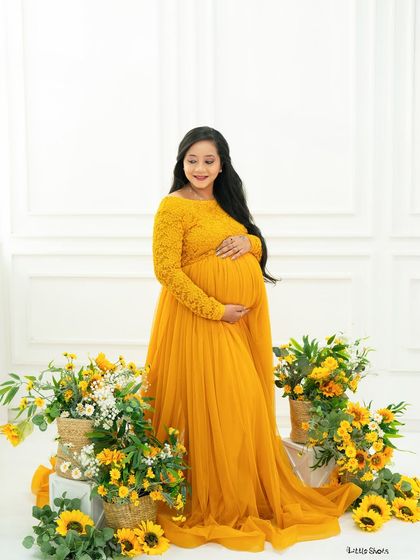 A radiant mother-to-be in a long-sleeved yellow lace gown. She stands surrounded by sunflowers, her hands on her bump, glowing with happiness.
