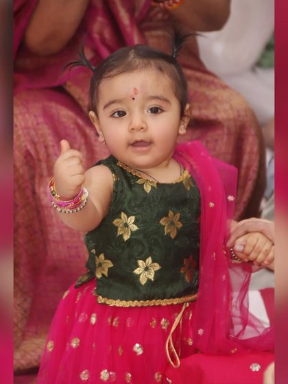 A baby girl in a traditional lehenga gives a thumbs-up, a perfect mix of cute and celebratory.