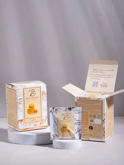 A clean, minimalist studio shot of Taste & Beyond cookie packaging on white pedestals. This e-commerce style focuses on the product and its unboxing experience.