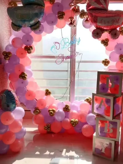 A beautiful baby shower or welcome home setup by a window, featuring a half-arch of pink, purple, and orange balloons, and transparent 'BABY' boxes filled with matching balloons.
