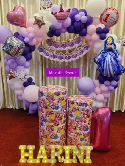 A royal first birthday celebration with a princess theme. The circular balloon arch in purple and pink is accented with a princess crown, a "Happy Birthday" banner, and themed plinths.