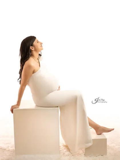 A full-length portrait showing the expecting mother seated on white blocks. Her upward gaze and the simple white gown create a feeling of hope and peacefulness.