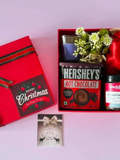 A festive Christmas hamper in a red box. It includes Hershey's hot chocolate, almond cookies, a scented candle, and a potli of treats.
