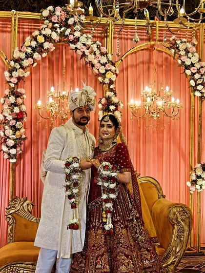 This is a final wedding stage setup during the Jaimala ceremony. The decor includes golden arches with white and pink floral arrangements, warm chandelier lighting, and a comfortable sofa for the couple.