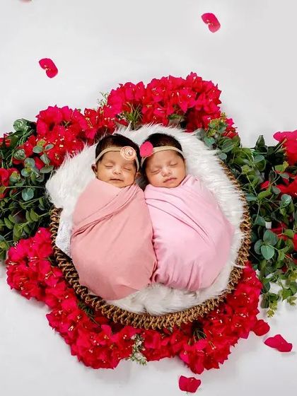 A beautiful floral-themed setup for newborn twins. Swaddled in pink, they are nestled together in a basket surrounded by vibrant red flowers.