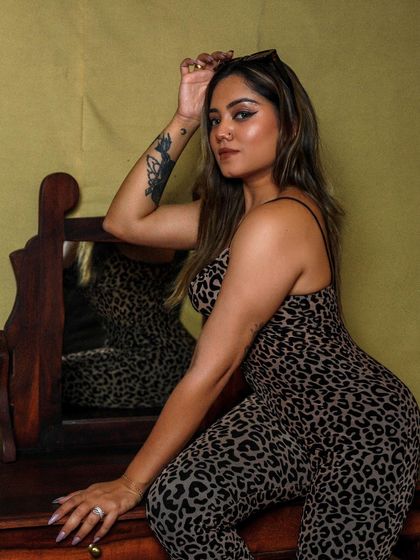 An indoor shot of a model in a leopard-print jumpsuit, posing confidently by a wooden vanity.