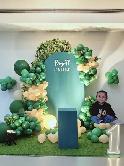 A "Wild One" first birthday setup for Rayirth, featuring a lush green backdrop and an organic balloon garland. A personalized cutout of the birthday boy adds a sweet, custom touch to the celebration.