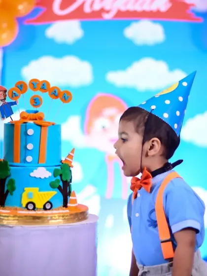 A little boy in a Blippi-inspired outfit, looking with excitement at his birthday cake.