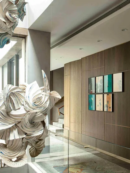 A close-up view of the hanging origami sculpture, showcasing the detailed spray painting and curved folding techniques. The artwork is juxtaposed with wood-paneled walls and a gallery of smaller art pieces, creating a rich, textural corridor.