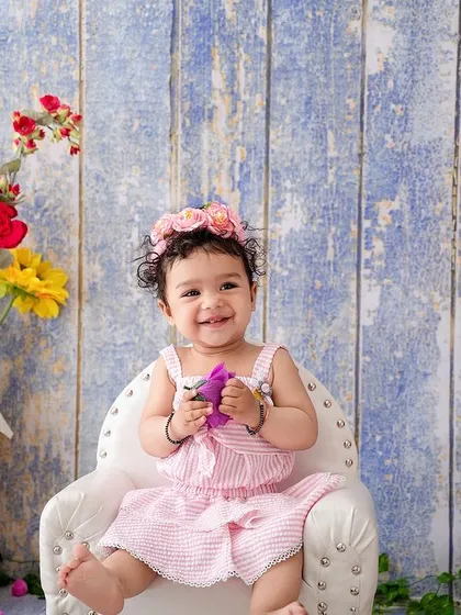A happy baby girl in a pink-striped outfit, sitting in a mini armchair against a rustic blue wooden backdrop.