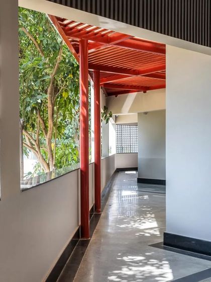 An open corridor at L'Atelier connects the interior spaces to the surrounding trees. The red structural elements and slatted ceiling create a rhythmic transition between the built form and the natural environment.