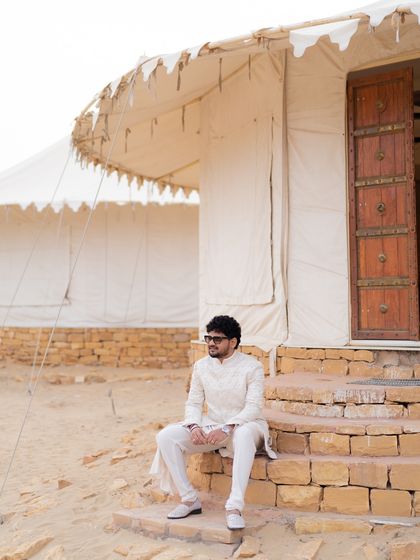 The groom looking cool and relaxed, seated outside a desert tent in his coordinated ivory outfit.