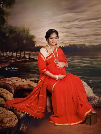 A beautiful, painterly portrait of a mother-to-be in a red saree, seated by a riverbank. The style is reminiscent of classic Indian art, creating a timeless and soulful image.