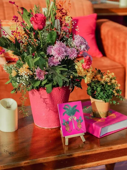 A vibrant centerpiece with a mix of fresh flowers in a bright pink pot, paired with a miniature easel holding a tropical-themed art card. It's all about those personal, quirky touches.