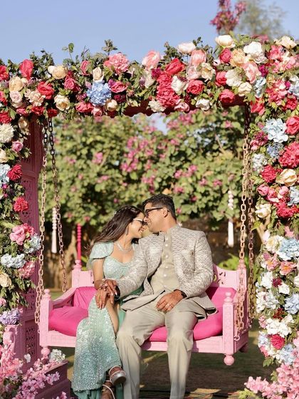 A floral-draped swing provides the perfect romantic spot for the couple at their pink Rajasthani Mehendi. The swing is framed by a lush arch of pink, red, and blue flowers, creating a beautiful photo backdrop.