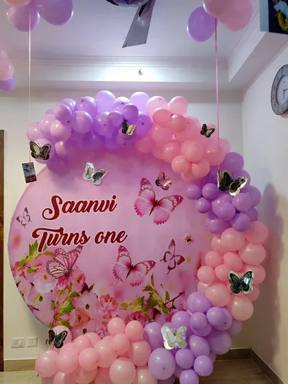 A close-up of a personalized butterfly-themed backdrop for Saanvi's first birthday. The custom-printed backdrop is framed with a pink and purple balloon garland for a beautiful photo spot.