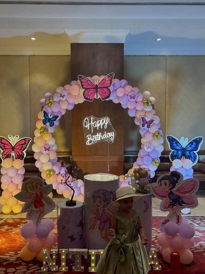 A butterfly and fairy theme for a third birthday, set up in a banquet hall. The design features a large balloon ring, character cutouts of fairies, butterfly accents, and a light-up number '3'.