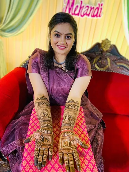 A happy bride showing her detailed wedding mehndi. The design includes personalized initials 'P' and 'R' on the palms, set within a traditional Rajasthani jharokha (window) frame, making it unique to her love story.