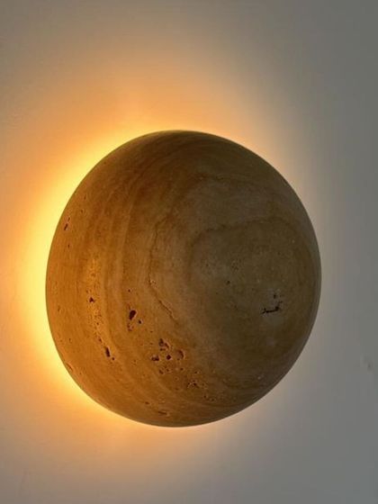 The travertine sphere wall light shown in a darker setting, creating a warm, moon-like effect on the wall.