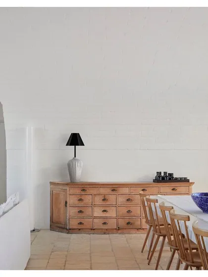 A table lamp from the 'Bernard and friends' collection sits atop a rustic wooden credenza. The lamp's simple, elegant form and natural wood texture bring a sense of calm and craftsmanship to the dining space.