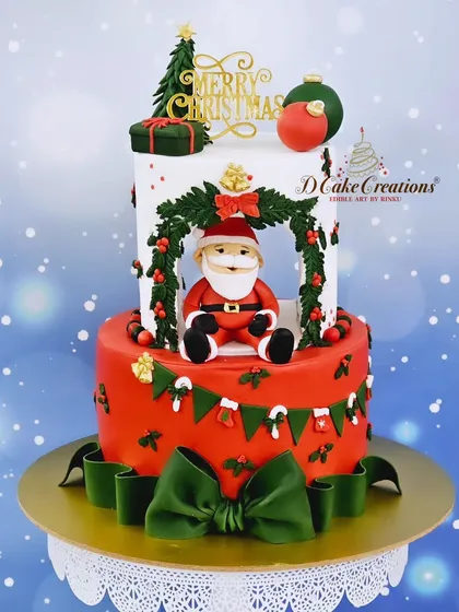 A two-tier Christmas cake with Santa Claus sitting in a festive wreath. The design includes a garland, bows, and candy canes.