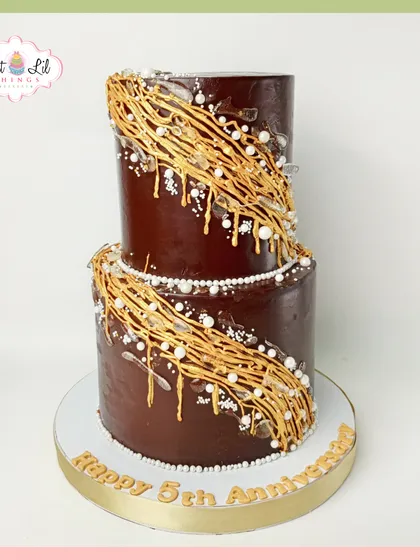 Indulge in pure luxury with this two-tiered chocolate anniversary cake. Draped in a rich chocolate ganache and adorned with intricate golden sugar work and edible pearls, it's a decadent and elegant way to celebrate your love story.