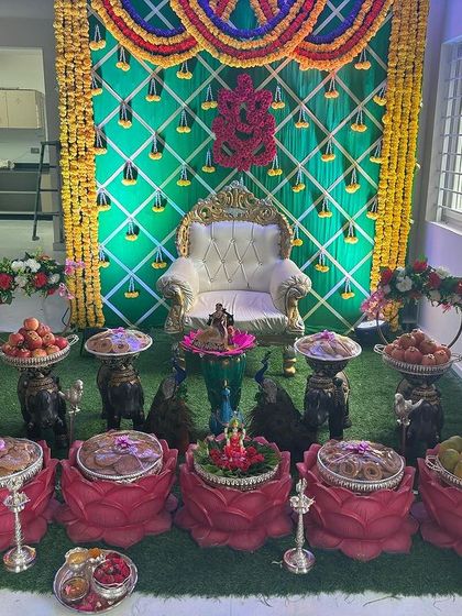 A vibrant decor setup for a home ceremony. The design includes a green backdrop with a Ganesha motif, yellow marigold garlands, and an offering table with lotus-shaped bowls.