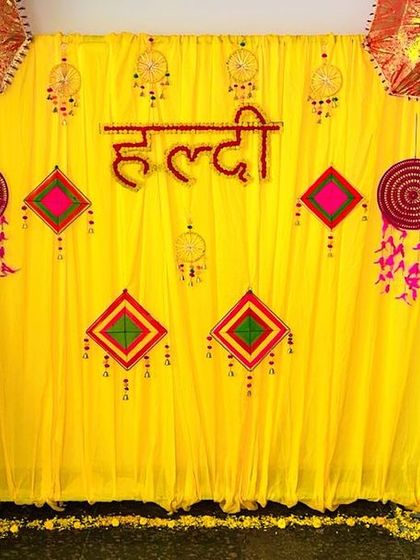 A closer look at our indoor Haldi setup. The "Haldi" lettering is made from red flowers, standing out beautifully against the yellow drapes, and is complemented by traditional Rajasthani hangings.