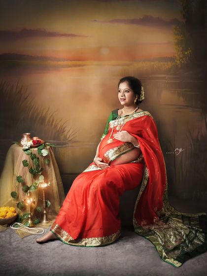 A serene and regal portrait. The mom-to-be is seated gracefully, cradling her baby bump, against a beautiful sunset backdrop that enhances the traditional and artistic feel of the shoot.