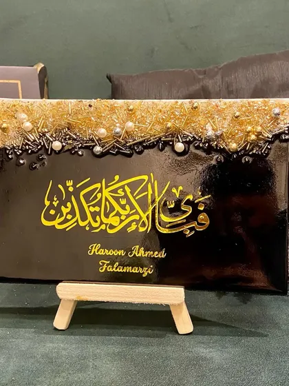 This personalized plaque features the verse "Fabi ayyi ala i rabbikuma tukazziban" in bold yellow calligraphy on a black background. The top is adorned with a textured mix of gold and silver embellishments, and it includes the client's name for a personal touch.