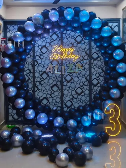 A chic and modern third birthday setup with a black and silver balloon circle, personalized neon sign, and a large light-up number '3'.