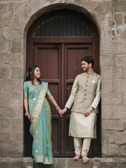 A classic couple portrait against a rustic stone wall and arched wooden door, showcasing a timeless pre-wedding look with traditional Indian attire.